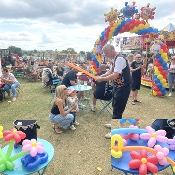 Children watching Magic Stuart twist a colourful balloon sword during an outdoor event